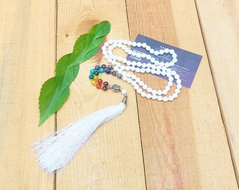 May include: A long beaded necklace featuring white spherical beads, a series of multi-colored chakra beads, a silver-toned metal charm depicting a face, and a long white fabric tassel. The necklace is arranged in a loop with green leaves.