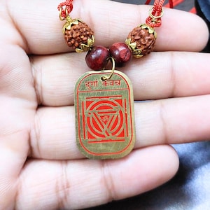May include: A gold-coloured pendant with red detailing and text, strung on a red cord with brown beads. The pendant is rectangular with a red geometric design and text at the top. The cord features two textured brown beads and two smaller red beads.