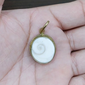 Puede incluir: Un colgante de oro con una concha blanca en su interior. La concha tiene un patrón en espiral.