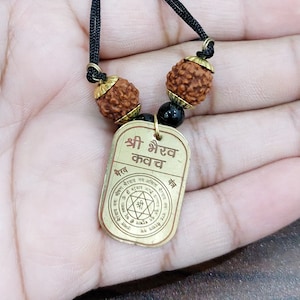 Puede incluir: Un collar con un colgante de oro, un cordón negro y cuentas marrones. El colgante es una placa dorada con el texto "श्री गरुव कवच" y un diseño circular con una estrella en el centro.