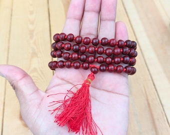 May include: A multi-strand bracelet composed of small, dark red wooden beads strung on a vibrant red cord. The bracelet features a prominent red tassel hanging from the center, secured with a small gold-colored metal band at the top of the fringe.