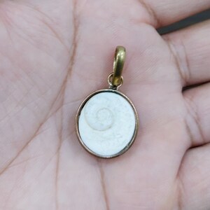 Puede incluir: Un colgante de concha blanca con un marco de latón. El colgante tiene forma ovalada y un patrón en espiral.