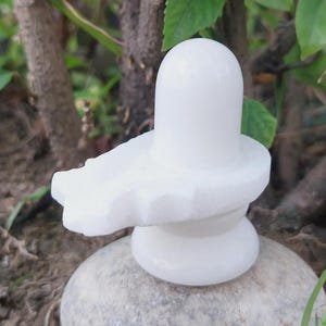 Puede incluir: Una estatua de Shiva Lingam de mármol blanco. La escultura presenta una base redondeada, una forma cilíndrica central y una plataforma en forma de pétalo. La estatua está sobre un fondo de follaje verde y una base de piedra natural.