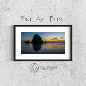 May include: A fine art print of a sunset over the ocean with Haystack Rock in the foreground. The rock is a dark green color and is reflected in the water. The sky is a mix of orange, pink, and purple.