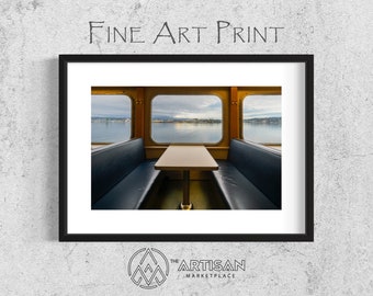 May include: A fine art print of a photograph of the interior of a ferry boat. The photo shows a wooden table with two benches facing each other. The windows of the ferry boat are visible in the background, showing a view of a body of water with a city skyline in the distance.