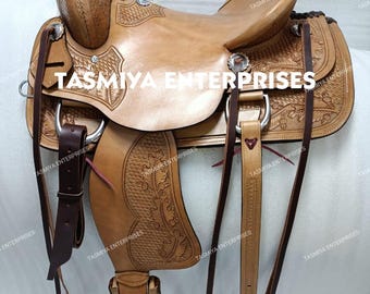 May include: A light brown leather saddle with intricate floral tooling and silver-toned hardware. The saddle features a high cantle, a horn, and stirrups. The text "TASMIYA ENTERPRISES" is visible on the saddle.