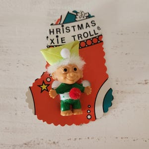 May include: A vintage Christmas Pixie Troll doll on a red stocking-shaped card. The troll has white hair, a green hat, and a red pom-pom. The card has the text "CHRISTMAS XIE TROLL".