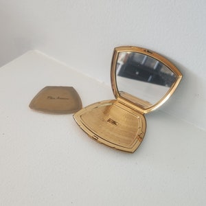 May include: A gold-toned compact mirror with a triangular shape. The mirror is open and the inside is lined with a gold-toned pattern. The back of the compact has the inscription "Elia Ancona" in black lettering.