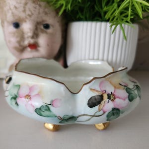 May include: A small, round, white porcelain bowl with a scalloped rim and three gold-painted feet. The exterior features hand-painted pink flowers, green leaves, and a bumblebee with black and yellow stripes. The rim is trimmed in gold.