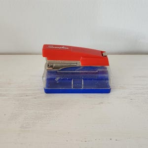 May include: A red and blue Swingline stapler. The stapler has a red top and a blue base with a clear plastic section. The word "Swingline" is printed in silver on the top. The stapler is on a white surface.