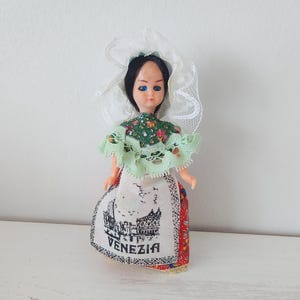 May include: A small doll in traditional Venetian attire. The doll has black hair, blue eyes, and a white lace veil. It wears a green floral top and a skirt with a "VENEZIA" graphic and a cityscape illustration.