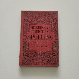 May include: A vintage, red book titled "Progressive Course in Spelling, Part One" by J.N. Hunt. The cover features ornate floral and scrollwork designs. The book is slightly worn, suggesting age and use.