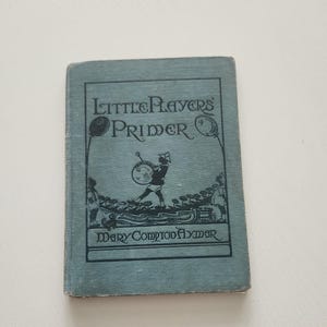 May include: A vintage book titled "Little Players Primer" by Mary Compton Aymar. The cover is a faded blue with black lettering and illustrations. The book is rectangular and shows signs of wear.