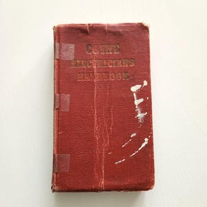 May include: A vintage red book with gold lettering that reads "COYNE ELECTRICIANS HANDBOOK." The book has visible wear and tear, including scratches and tape. The book is a rectangular shape.