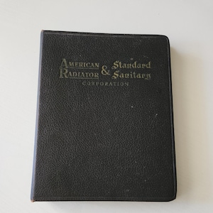 May include: A rectangular, black, textured-cover book with the gold-embossed text American Radiator & Standard Sanitary Corporation on the front cover. The book has a hard, grained exterior finish and a simple, clean, rectangular shape.