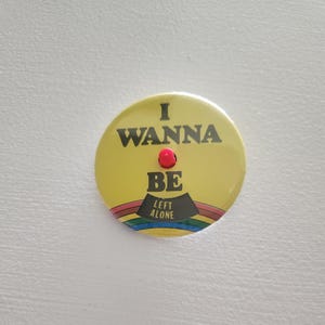 May include: A yellow button with the text "I WANNA BE LEFT ALONE" in black. A red dot is above the word "BE", and a rainbow design is at the bottom. The button is round.