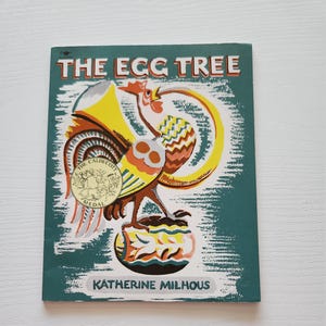 May include: A vintage children's book titled "The Egg Tree" with a teal cover. The cover features a colorful illustration of a rooster and a gold Caldecott Medal. The author's name, Katherine Milhous, is printed at the bottom.