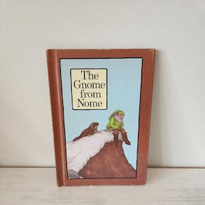 May include: A vintage children's book titled "The Gnome from Nome." The cover features an illustration of a gnome in a green outfit, a brown mountain, and the text in a white box against a light blue background. The book has a brown border.
