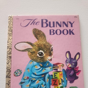 May include: A vintage children's book titled "The Bunny Book" with a pink cover. The cover features a cartoon bunny wearing blue pajamas with carrots printed on them. The bunny is playing with alphabet blocks and a purple bunny is standing next to it. The book is a "Little Golden Book".