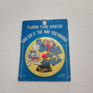 May include: A vintage button with the text "LOVE for SALE" in blue and yellow, with a red heart and a cartoon figure. The button is attached to a blue card with the text "TURN THE KNOB AND SAY IT THE WAY YOU WANNA".