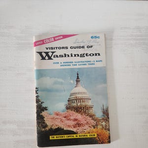 May include: A vintage "Visitors Guide of Washington" travel booklet. The cover features a color illustration of the U.S. Capitol building with pink cherry blossoms. The text includes "Latest Color Edition" and "The Nation's Capital in Natural Color."