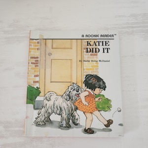May include: A children's book titled "KATIE DID IT" by Becky Bring McDaniel. The cover illustration depicts a young girl in an orange dress kicking a ball, with a fluffy white dog nearby. The book is part of the "A Rookie Reader" series.
