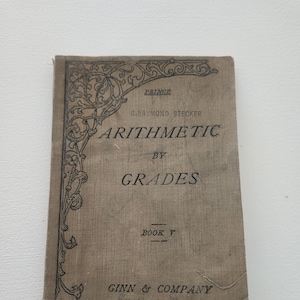May include: A vintage book titled "Arithmetic by Grades, Book V" by C. Raymond Stecker. The book cover is brown with a decorative black border and the text is printed in black.