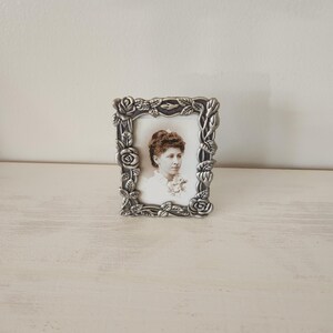 May include: A small, rectangular, silver-colored picture frame with a floral design. The frame holds a black and white portrait of a woman. The frame is resting on a light-colored surface against a white wall.