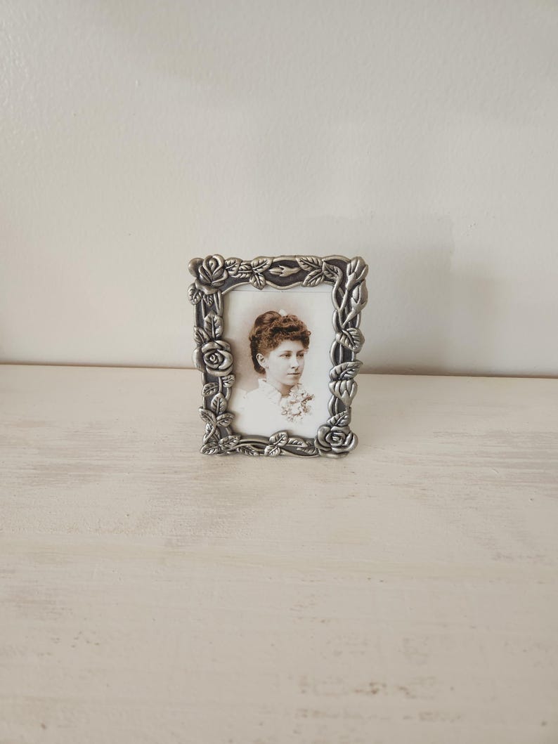 May include: A small, rectangular, silver-colored picture frame with a floral design. The frame holds a black and white portrait of a woman. The frame is resting on a light-colored surface against a white wall.