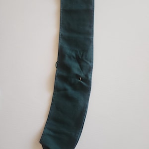 May include: A dark green fabric sash, possibly for a garment or accessory, lies flat. A pin is inserted near the center. The sash is rectangular with a pointed end and a folded top edge.