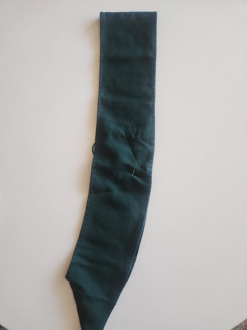 May include: A dark green fabric sash, possibly for a garment or accessory, lies flat. A pin is inserted near the center. The sash is rectangular with a pointed end and a folded top edge.