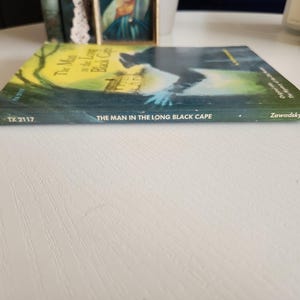 Pode incluir: Um livro intitulado "The Man in the Long Black Cape" de Zawadsky. A capa do livro apresenta uma silhueta escura de uma pessoa com um longo manto preto em p&eacute; em frente a uma casa.