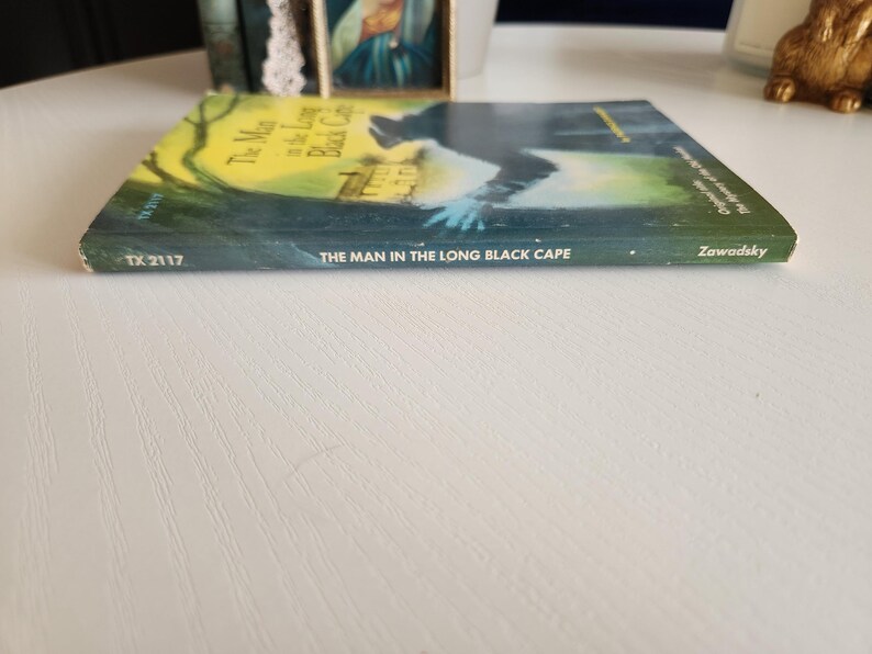 Pode incluir: Um livro intitulado "The Man in the Long Black Cape" de Zawadsky. A capa do livro apresenta uma silhueta escura de uma pessoa com um longo manto preto em p&eacute; em frente a uma casa.