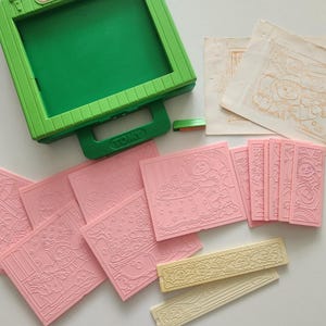 May include: A green and pink vintage drawing toy set. The set includes a green drawing board, a green eraser, pink stamps with various designs, and two off-white stampers. The stamps feature images of a bear, a girl, and other designs.