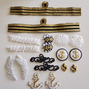 May include: Assortment of decorative patches and appliques. Includes gold and black striped bands, white embroidered swirls, bee patches, monogrammed patches, silver wing appliques, black lace appliques, anchor patches, and gold anchor pins.