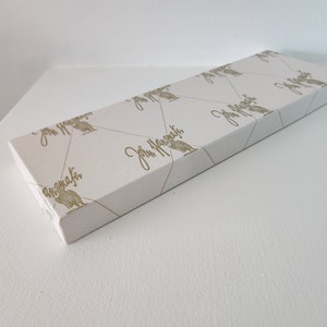 May include: A white rectangular box with a gold pattern of birds and the text "John Lewis" repeated across the surface. The box is closed and sitting on a white surface.