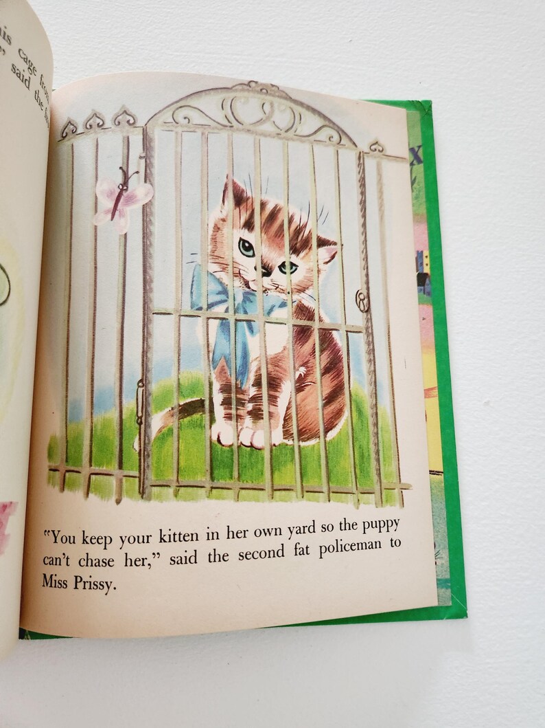 Może przedstawiać: Ilustracja z książki dla dzieci przedstawiająca brązowo-białego kociaka z niebieskimi oczami, noszącego niebieską kokardkę, patrzącego przez białą metalową bramkę. Tekst brzmi: "Trzymasz swojego kociaka na swoim podw&oacute;rku, żeby szczeniak nie m&oacute;gł go gonić", powiedział drugi gruby policjant do Miss Prissy.