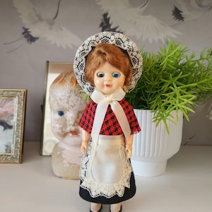 May include: A vintage doll with red hair, blue eyes, and a white lace bonnet. The doll wears a red and black checkered top, a white apron with lace trim, and a black skirt. The doll is standing on a white shelf.
