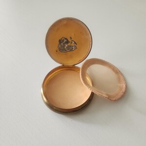 May include: A round gold-toned metal powder compact with an open hinged lid. The interior features a light peach-colored powder base and a separate circular mesh sifter with a beige fabric trim resting beside the open container on the surface.