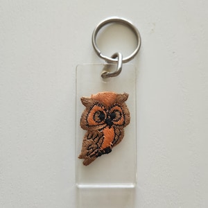 May include: A clear rectangular keychain with a metal ring. The keychain features an embroidered owl design in shades of brown and orange. The owl is centered on the clear plastic.