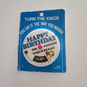May include: A vintage birthday button with a blue background and the text "TURN THE KNOB AND SAY IT THE WAY YOU WANNA." The button reads "HAPPY BIRTHDAY FOR SOMEONE WHO IS REALLY FLAKY."