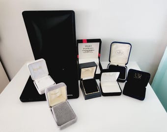 May include: A collection of eight black, gray, and blue velvet ring boxes. The boxes are all different sizes and shapes. Some of the boxes have a white interior, while others have a black interior. The boxes are all closed, and the lids are not open. The boxes are arranged in a row on a white surface.