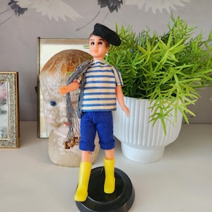 May include: A vintage doll dressed in a sailor outfit. The doll wears a black beret, a blue and white striped shirt, blue shorts, and yellow boots. It stands on a black base. A decorative plant and a vintage doll head are in the background.