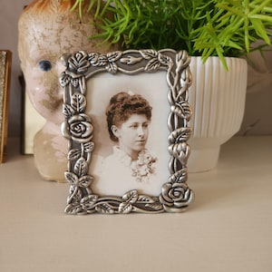 May include: A small, square, silver-colored picture frame with a portrait of a woman. The frame is decorated with roses and leaves. The portrait is black and white. The frame is sitting on a white surface.