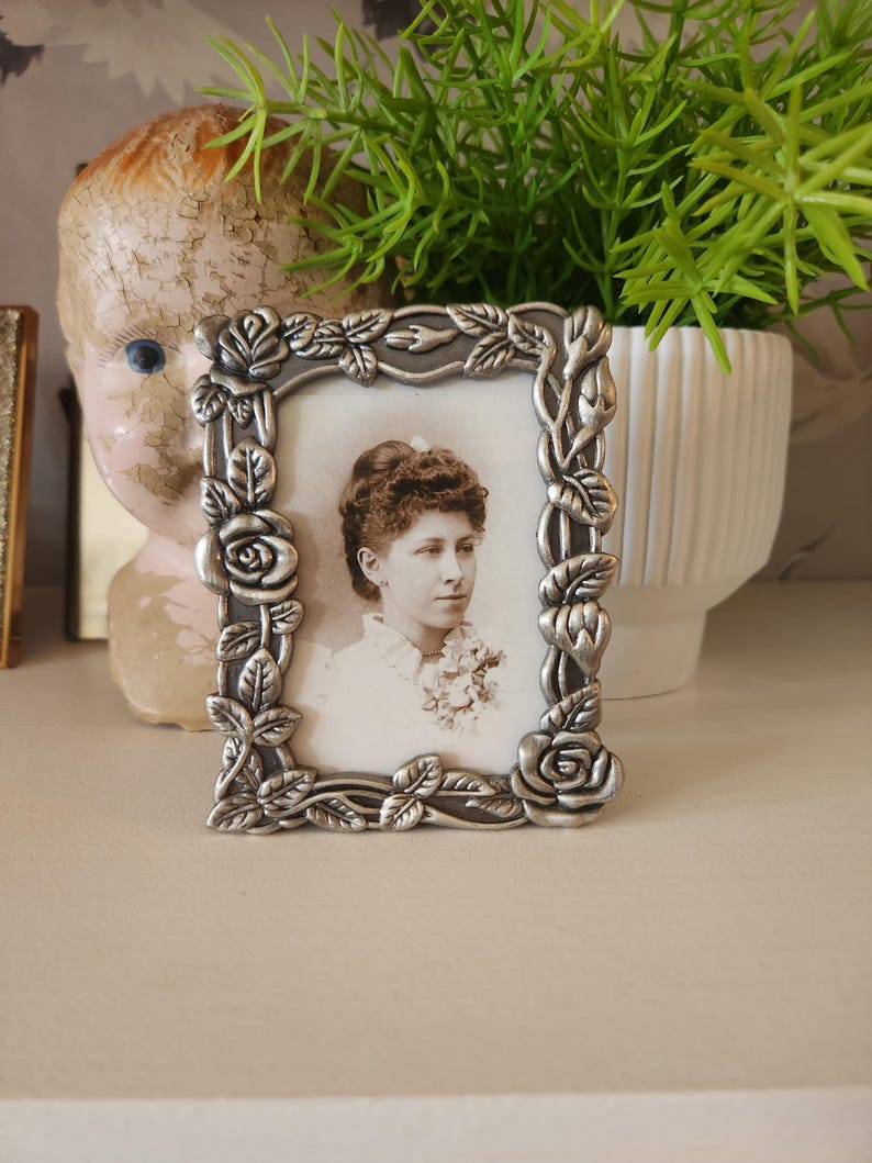 May include: A small, square, silver-colored picture frame with a portrait of a woman. The frame is decorated with roses and leaves. The portrait is black and white. The frame is sitting on a white surface.