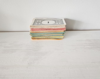 May include: A stack of rectangular paper game money featuring a white top bill with a black printed number one in a circular frame. The remaining bills in the stack display various pastel colors including pink, light blue, and yellow along the edges.