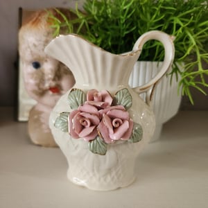 May include: A cream-colored ceramic pitcher with a handle and a spout, adorned with three pink rose embellishments and green leaves. The pitcher has a gold trim and a textured surface. Decorative item.
