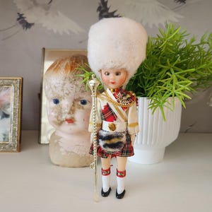 May include: A vintage doll dressed in a Scottish military uniform, complete with a white fur hat, red and black plaid kilt, and a gold staff. The doll has a painted face and is standing on a white surface.