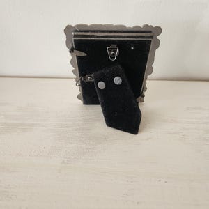 May include: A small, ornate silver-colored picture frame with a black velvet backing. The frame has a decorative, scalloped edge. A black easel-style stand is attached to the back, allowing the frame to stand upright. The frame is designed to hold a small photograph.