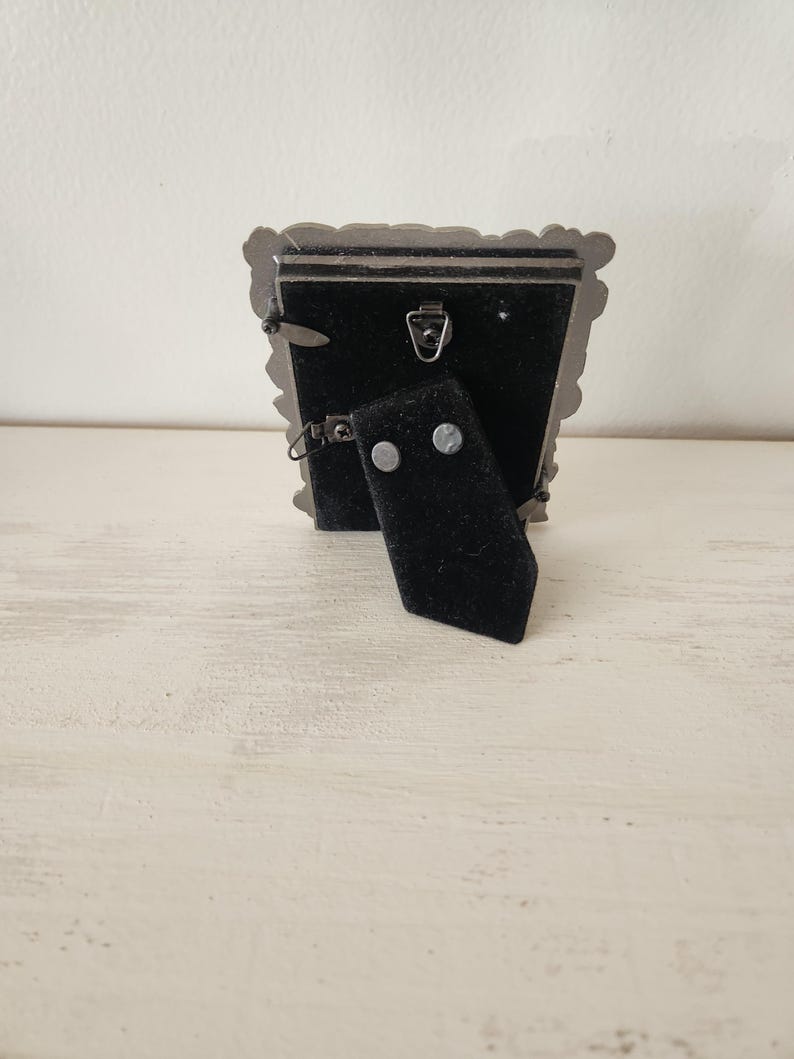 May include: A small, ornate silver-colored picture frame with a black velvet backing. The frame has a decorative, scalloped edge. A black easel-style stand is attached to the back, allowing the frame to stand upright. The frame is designed to hold a small photograph.